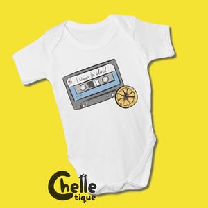 Könnte beinhalten: Weißer Baby-Body mit einem Kassetten-Motiv und einer Zitronenscheibe. Auf der Kassette steht der Text "I Wanna be adored". Der Body liegt vor gelbem Hintergrund.