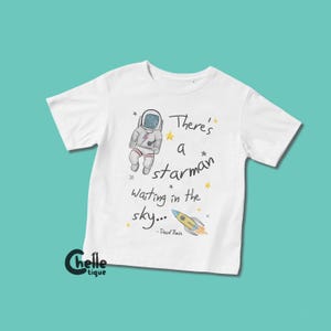 Pode incluir: T-shirt branca com um astronauta de desenho animado, estrelas e um foguete. O texto "There's a starman waiting in the sky..." está impresso na camiseta, com "- David Bowie" abaixo. A camiseta tem mangas curtas.