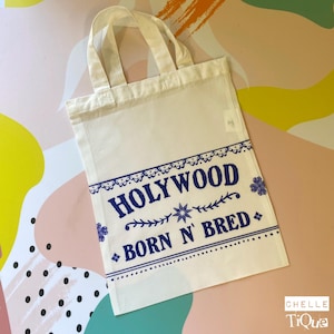 Könnte beinhalten: Eine weiße Canvas-Tragetasche mit blauem Text "HOLLYWOOD BORN N' BRED". Die Tasche hat ein rechteckiges Design, einen Griff und einen dekorativen blauen Rand.