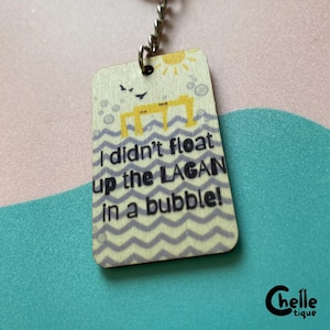 Op de afbeelding: Een rechthoekige houten sleutelhanger met een gele brug-afbeelding, een zon en vogels boven een grijs golfpatroon. De tekst luidt: I didn't float up the LAGAN in a bubble! De sleutelhanger is bevestigd aan een zilverkleurige metalen ketting en ring.