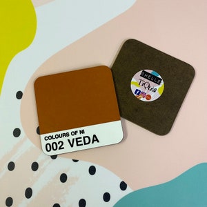 May include: A brown square coaster with the text "Colours of NI 002 Veda" printed on it. The coaster is sitting on a white surface with black polka dots.