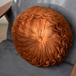 May include: A round brown velvet cushion with a tufted centre and a braided edge. The cushion is sitting on a grey chair.