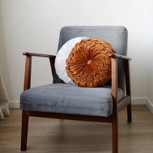May include: A grey velvet armchair with a wooden frame and a round, tufted cushion in shades of white and orange.
