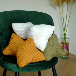 Op de afbeelding: Drie stervormige kussens in een groene fluwelen stoel. De kussens zijn wit, mosterdgeel en olijfgroen.