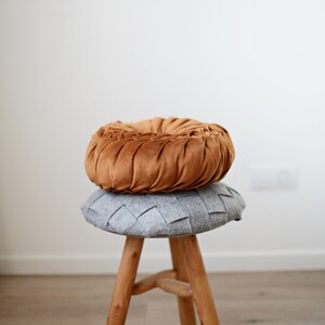 May include: A wooden stool with three legs, topped with a grey woven cushion and a brown velvet cushion.
