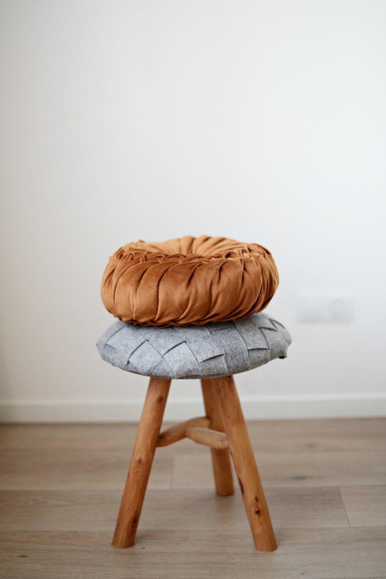 May include: A wooden stool with three legs, topped with a grey woven cushion and a brown velvet cushion.