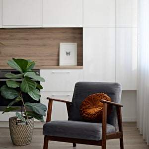May include: A grey armchair with wooden legs and a round, orange velvet cushion. The chair is in a modern living room with white cabinets and a potted plant.