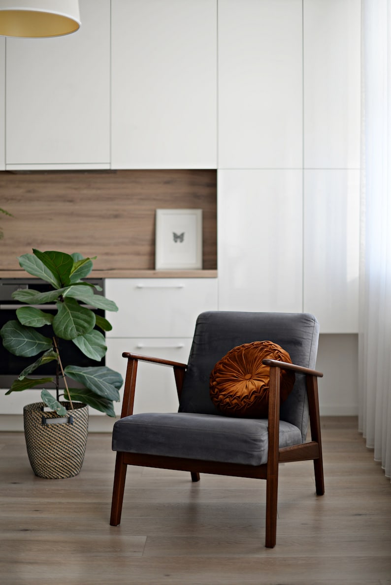 May include: A grey armchair with wooden legs and a round, orange velvet cushion. The chair is in a modern living room with white cabinets and a potted plant.
