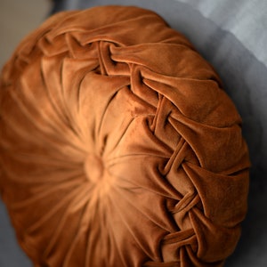 May include: A round, brown velvet cushion with a tufted design. The cushion has a button in the centre and is surrounded by a braided pattern.