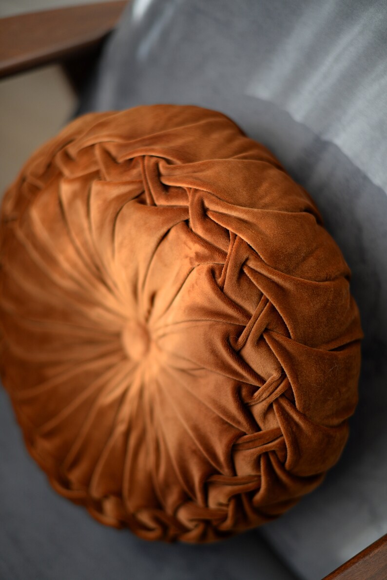 May include: A round, brown velvet cushion with a tufted design. The cushion has a button in the centre and is surrounded by a braided pattern.