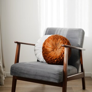 May include: A grey velvet armchair with wooden legs and a brown and white round cushion. The chair is in a room with white walls and a white curtain.