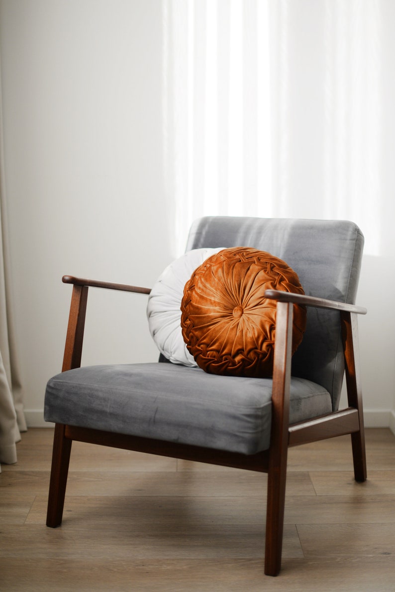 May include: A grey velvet armchair with wooden legs and a brown and white round cushion. The chair is in a room with white walls and a white curtain.