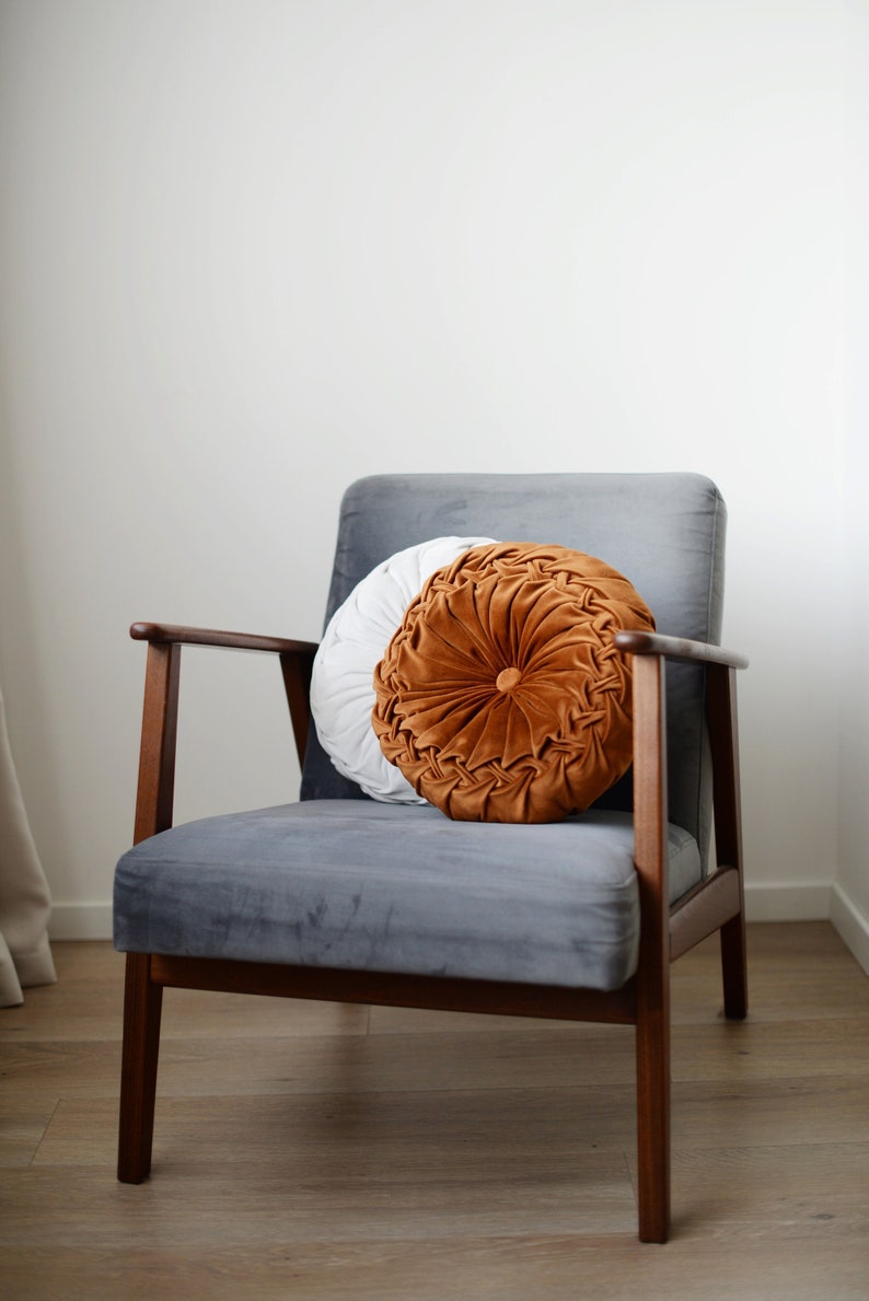 May include: A grey velvet armchair with a wooden frame and a round, tufted cushion in shades of white and orange.