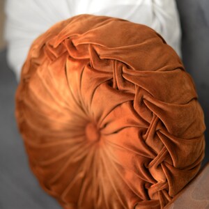 May include: A round, orange velvet cushion with a button in the centre and a tufted design. The cushion is on a grey surface.