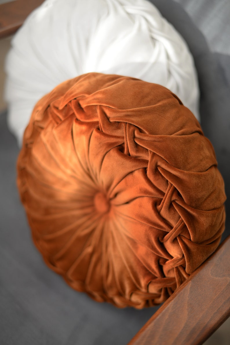 May include: A round, orange velvet cushion with a button in the centre and a tufted design. The cushion is on a grey surface.