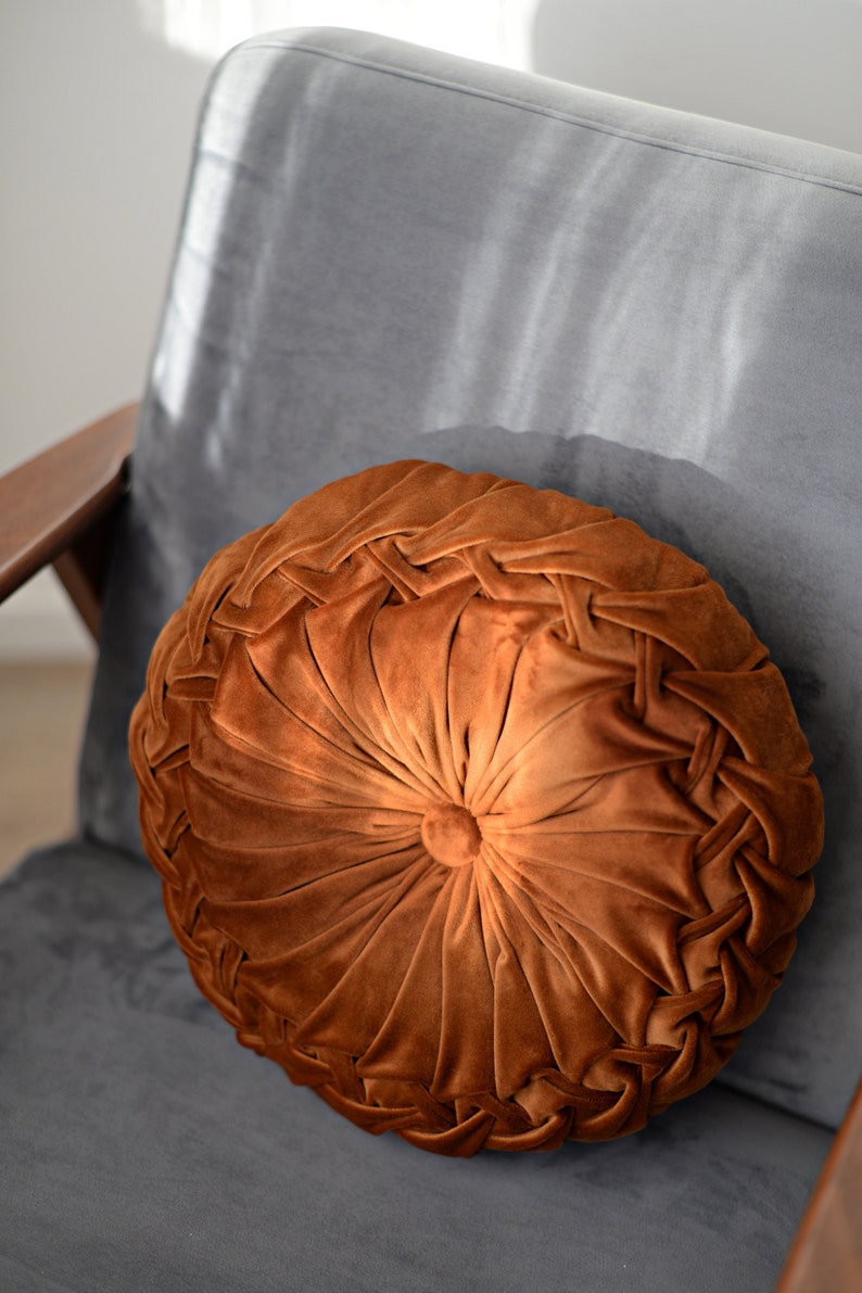 May include: A round brown velvet cushion with a tufted centre and a braided edge. The cushion is sitting on a grey chair.