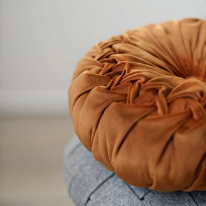 May include: A round, brown velvet cushion with a tufted design. The cushion is sitting on top of a grey cushion.