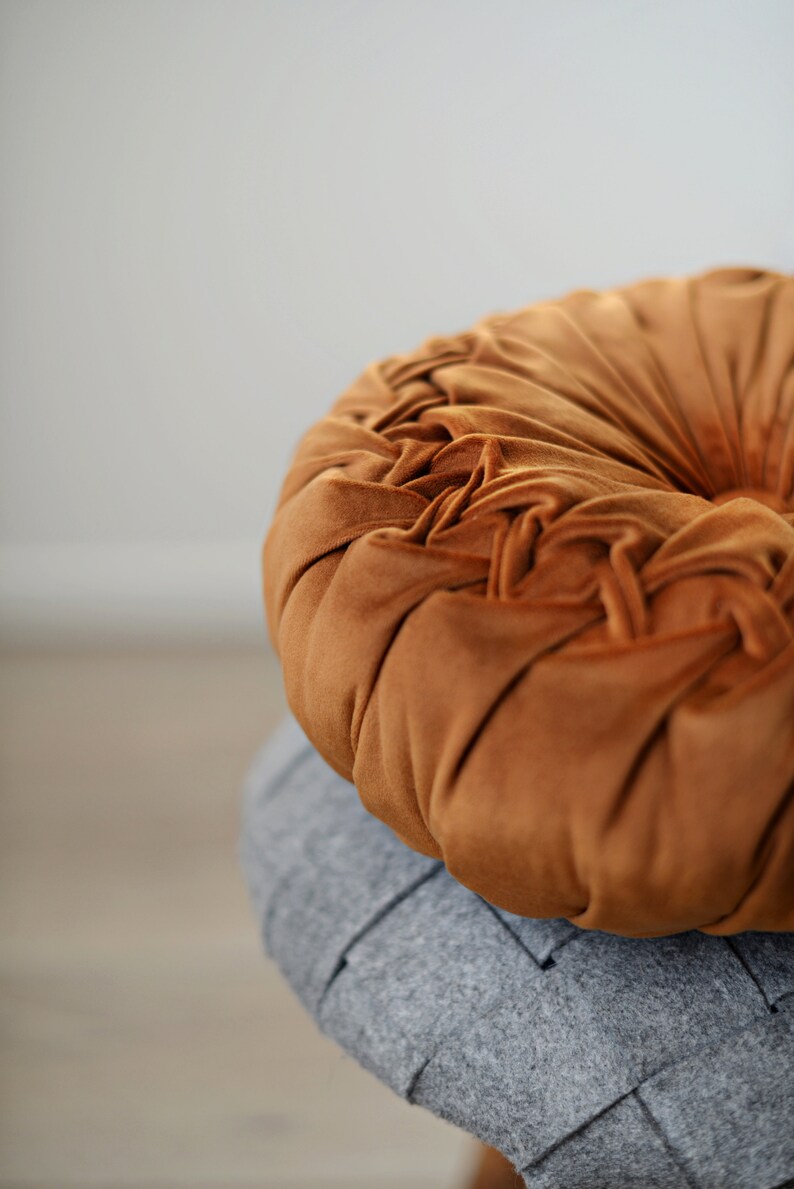 May include: A round, brown velvet cushion with a tufted design. The cushion is sitting on top of a grey cushion.