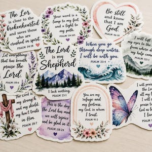 May include: A collection of eleven die-cut stickers featuring various Bible verses and religious themes. Each sticker displays unique watercolor illustrations including floral wreaths, mountain landscapes, ocean waves, a butterfly, and a wooden cross.