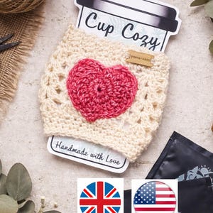 Puede incluir: Un portavasos de ganchillo color crema con un aplique de corazón rojo. El portavasos está envuelto alrededor de una taza de papel con las palabras "Cup Cozy" impresas. Una pequeña etiqueta de madera dice "yamnyrobin". Las palabras "Handmade with Love" están impresas en una etiqueta.