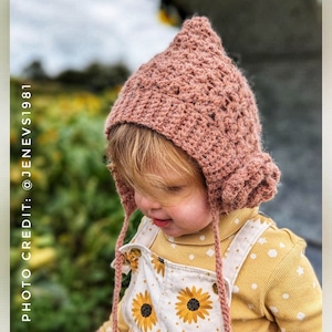 Op de afbeelding: Een klein kind draagt ​​een roze gebreide muts met een puntige top en bandjes. De muts heeft een textuurontwerp en wordt gedragen over een geel shirt met witte stippen en een witte overall met een zonnebloemmotief.