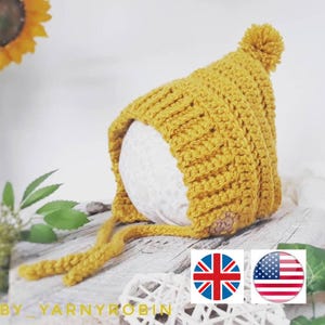 Può includere: Un berretto per neonato in crochet giallo con un pon pon in cima. Il berretto ha un laccio che può essere legato sotto il mento.