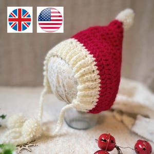 May include: A handmade crochet hat in red and cream. The hat has a pointed top with a cream pom-pom and ear flaps with cream pom-poms. The hat is likely for a baby or small child.