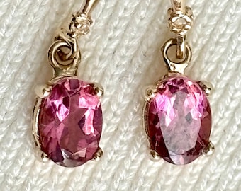 May include: A pair of gold earrings with pink oval-shaped gemstones. The earrings have a simple design with a small gold loop at the top and a dangling gemstone.