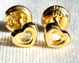 May include: A pair of gold-colored heart-shaped stud earrings. The earrings have a hollow heart design and are set on a round, textured backing. The earrings are displayed on a white, textured surface.