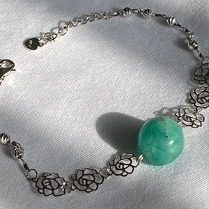 Op de afbeelding: Een zilveren armband met een groene edelsteen en zilveren roosvormige bedels.