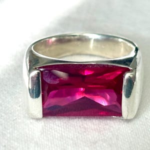 Peut inclure: Une bague en argent avec une grosse pierre précieuse rectangulaire de couleur magenta. La bague a un anneau épais et un design simple et moderne. La pierre précieuse est facettée, captant la lumière et créant un effet scintillant. La bague est posée sur un fond blanc.