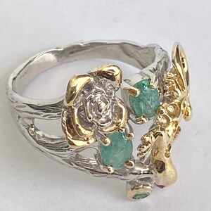 May include: A silver ring with a gold rose and green gemstones. The ring is designed to look like a branch with a bird perched on it.