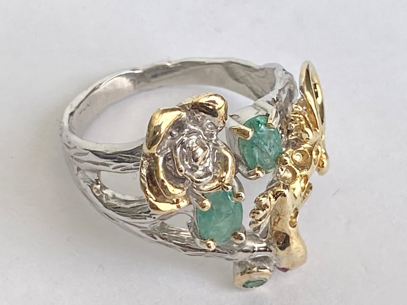 May include: A silver ring with a gold rose and green gemstones. The ring is designed to look like a branch with a bird perched on it.