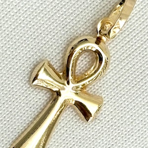 May include: A gold-colored Ankh pendant, a symbol of life, is displayed against a white background. The pendant features a loop at the top, a vertical bar, and a horizontal bar, all in a polished finish. The Ankh is a classic Egyptian symbol.