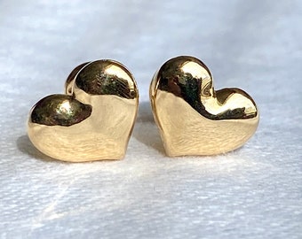 May include: A pair of polished gold heart-shaped stud earrings. The earrings have a smooth, reflective surface, catching the light and creating highlights. The heart shape is rounded and symmetrical, suggesting a classic design.