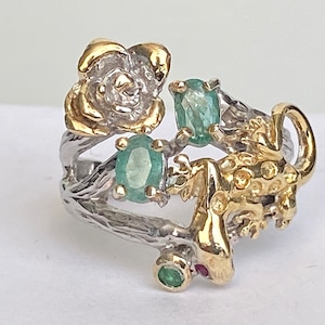 May include: A silver and gold ring with a detailed design of a flower, a lizard, and green gemstones.