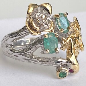 May include: A silver ring with a gold flower, a gold lizard, and green gemstones. The ring is designed to look like a branch with a flower and lizard on it.
