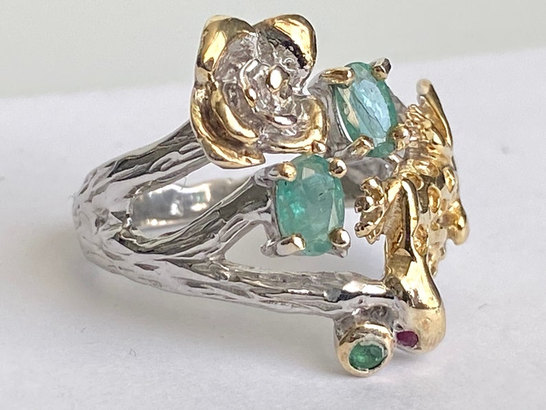 May include: A silver ring with a gold flower, a gold lizard, and green gemstones. The ring is designed to look like a branch with a flower and lizard on it.