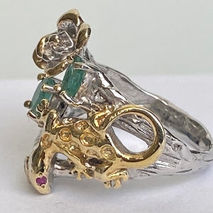 May include: A silver and gold ring with a green emerald gemstone and a lizard design. The ring is set on a textured band.