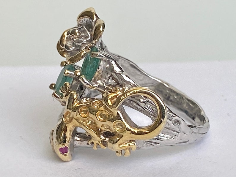 May include: A silver and gold ring with a green emerald gemstone and a lizard design. The ring is set on a textured band.