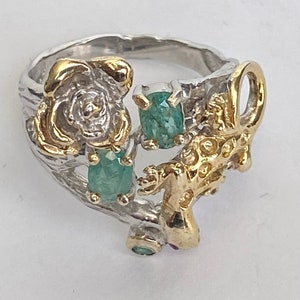 May include: A silver ring with a gold lizard design and green gemstones. The ring features a rose design with a gold center.