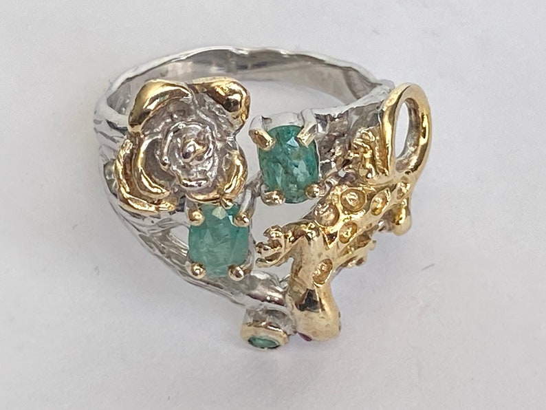 May include: A silver ring with a gold lizard design and green gemstones. The ring features a rose design with a gold center.
