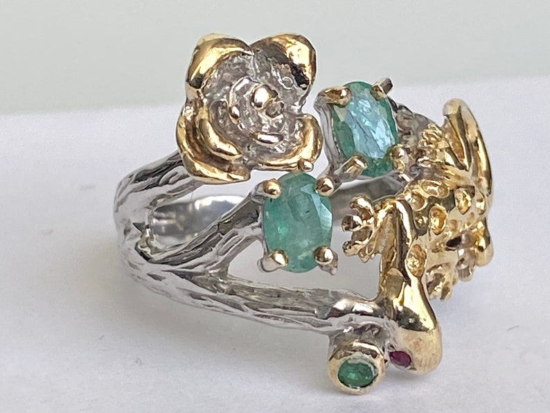 May include: A silver ring with a gold lizard, a gold flower, and green gemstones. The ring is designed to look like a branch.