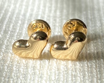 May include: A pair of gold heart-shaped stud earrings. The earrings have a polished finish and are set against a white, textured background. The earring posts are also gold.