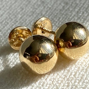 May include: Close-up of gold ball stud earrings. The earrings are highly polished and reflect the surrounding light. The earring posts and backs are also visible. The earrings are on a white, textured surface.
