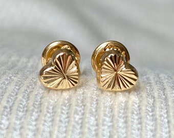 May include: A pair of gold-colored heart-shaped stud earrings. Each earring features a textured, radiating design on the heart's surface. The earrings have a post and backing for secure wear. The background is a soft, white fabric.