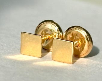 May include: A pair of gold-colored square stud earrings. The earrings have a simple, minimalist design with a polished finish. The posts and backs are also gold-colored. The earrings are displayed on a light-colored surface.