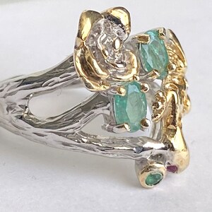 May include: A silver ring with a gold floral design and green gemstones. The ring is set on a white background.