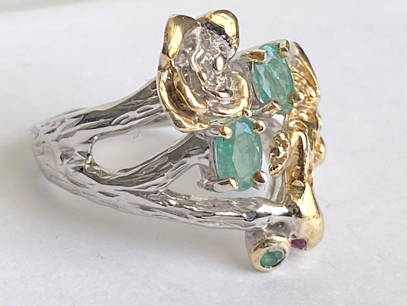 May include: A silver ring with a gold floral design and green gemstones. The ring is set on a white background.