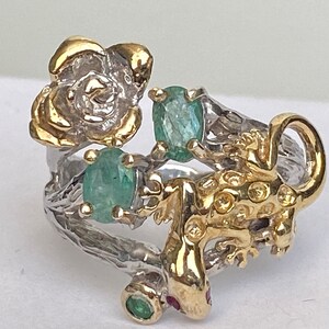 May include: A silver ring with a gold lizard, a gold flower, and green gemstones.